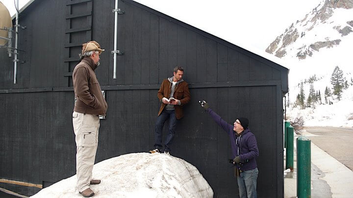 People preparing to photograph man standing on snow leaning against black barn