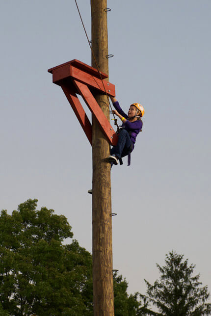 Harnessed person wearing yellow helmet climbing up pole